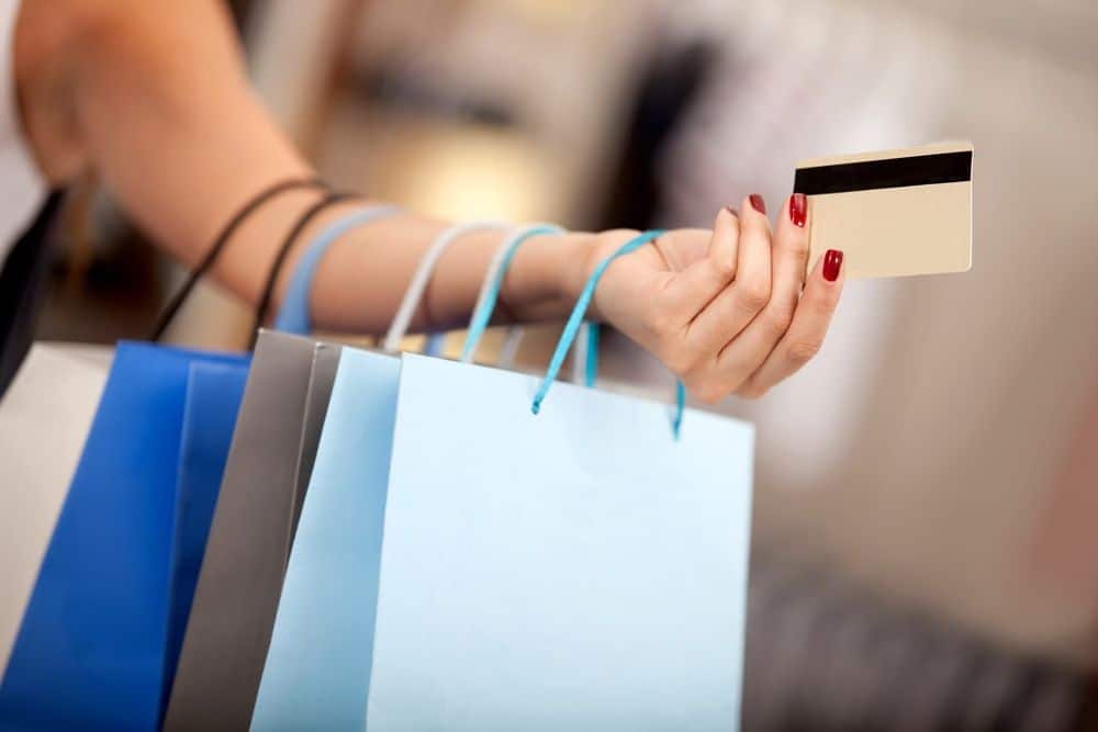 Woman shopping and paying with a debit or credit card A person with red-painted nails holds several shopping bags and a credit card, suggesting a shopping activity. The background is blurred, keeping the focus on the hand, bags, and card.