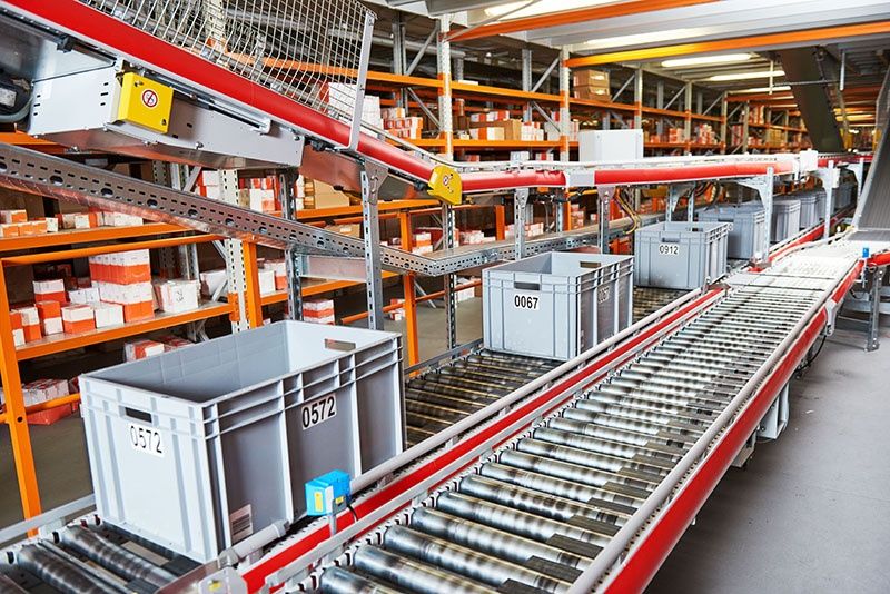 Plastic bins labeled with numbers move along a conveyor belt in a warehouse filled with shelves of orange and white packages. The setting appears organized and industrial, suggesting a logistics or distribution center.