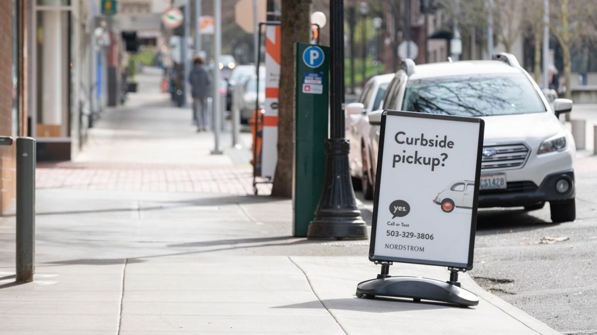 A sidewalk sign reads “Curbside pickup? yes” with a phone number and a small car graphic, promoting Nordstrom’s curbside service. Cars are parked on the street and pedestrians walk in the background.