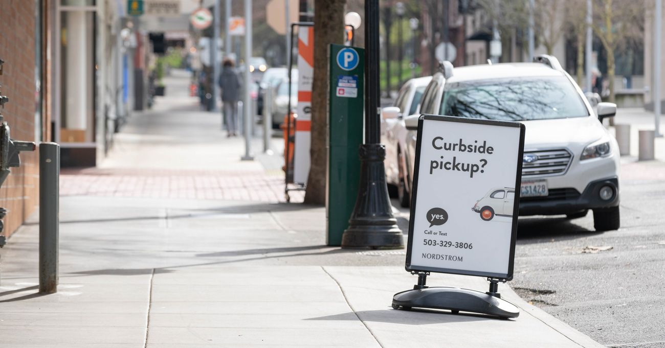A sidewalk sign reads “Curbside pickup? yes” with a phone number and a small car graphic, promoting Nordstrom’s curbside service. Cars are parked on the street and pedestrians walk in the background.