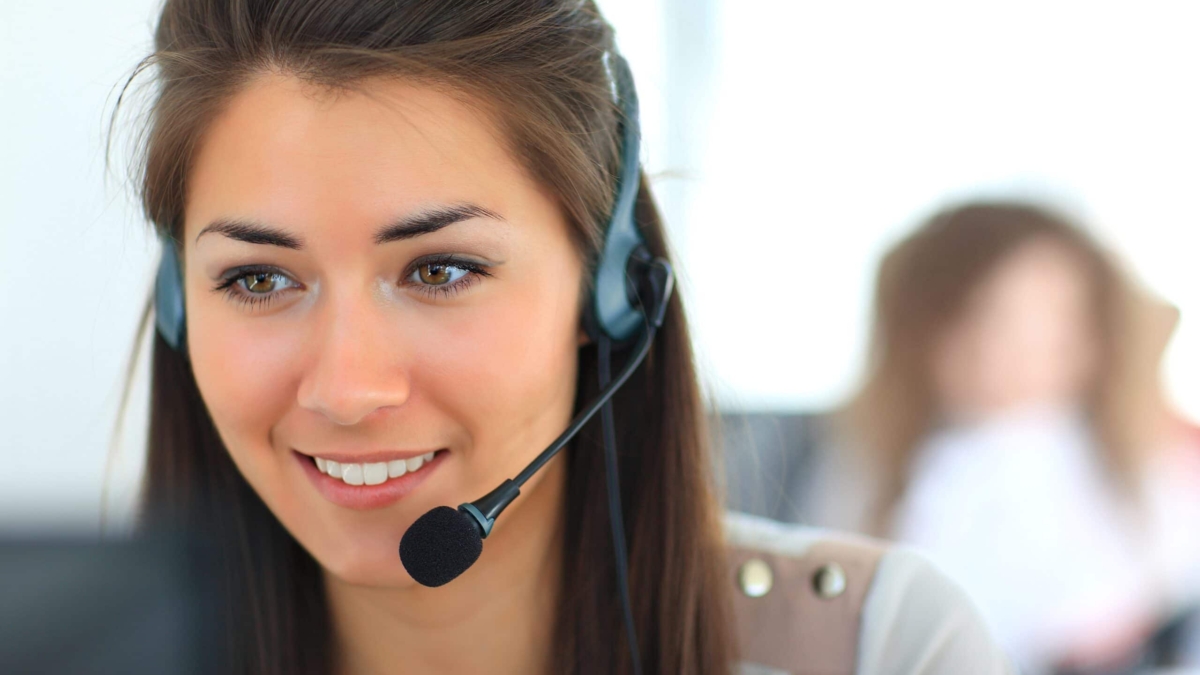 A woman with long brown hair wearing a headset smiles while working at a computer in an office. Another person is blurred in the background.