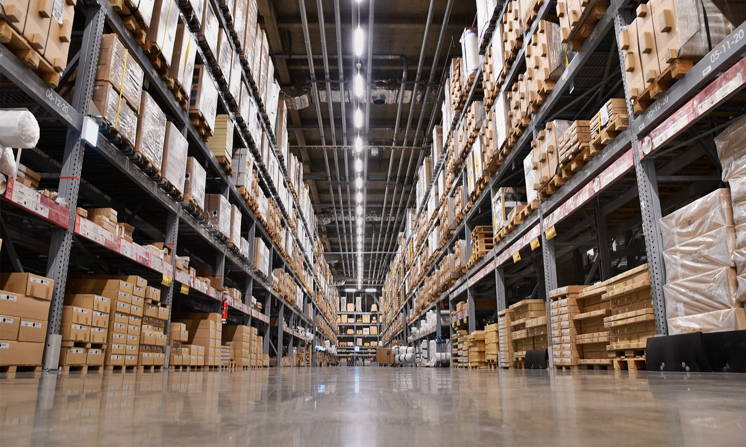 distribution-center-capacity-analysis-mobile View of a large warehouse with tall shelves stacked with cardboard boxes, ideal for warehouse capacity analysis, bright overhead lighting, and a shiny concrete floor, seen from a low angle between the aisles.
