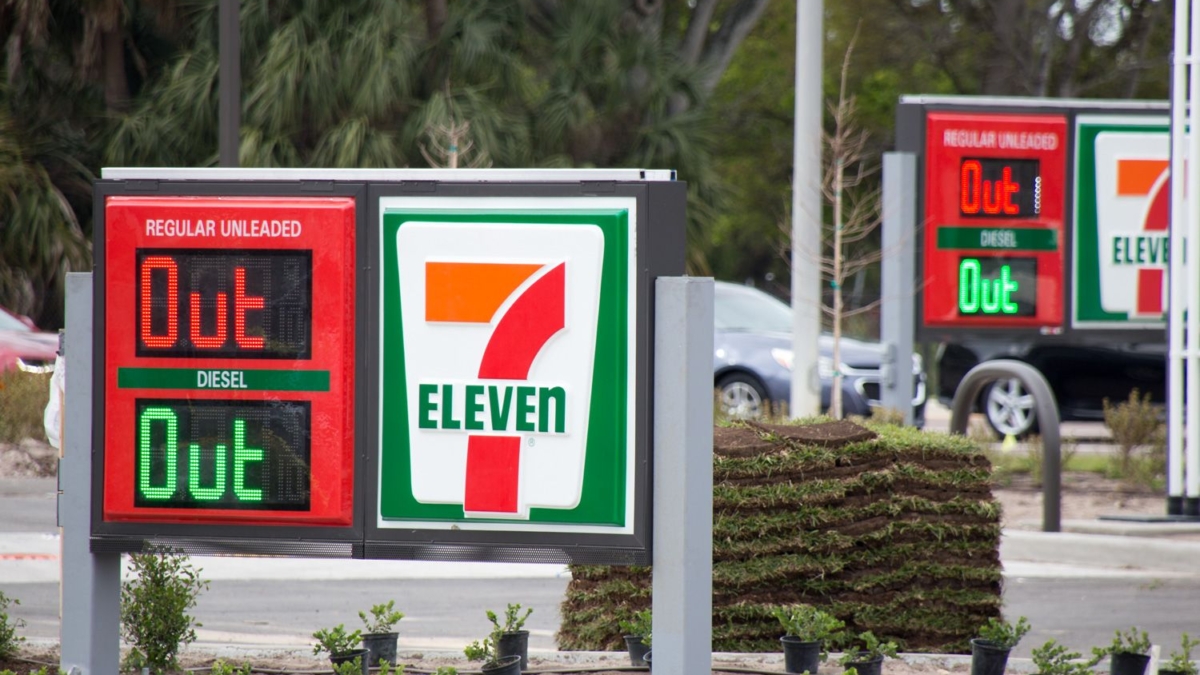 A 7-Eleven gas station sign displays OUT for both regular unleaded and diesel fuel, indicating they are unavailable. The same message appears on a second sign in the background. Small plants line the area.