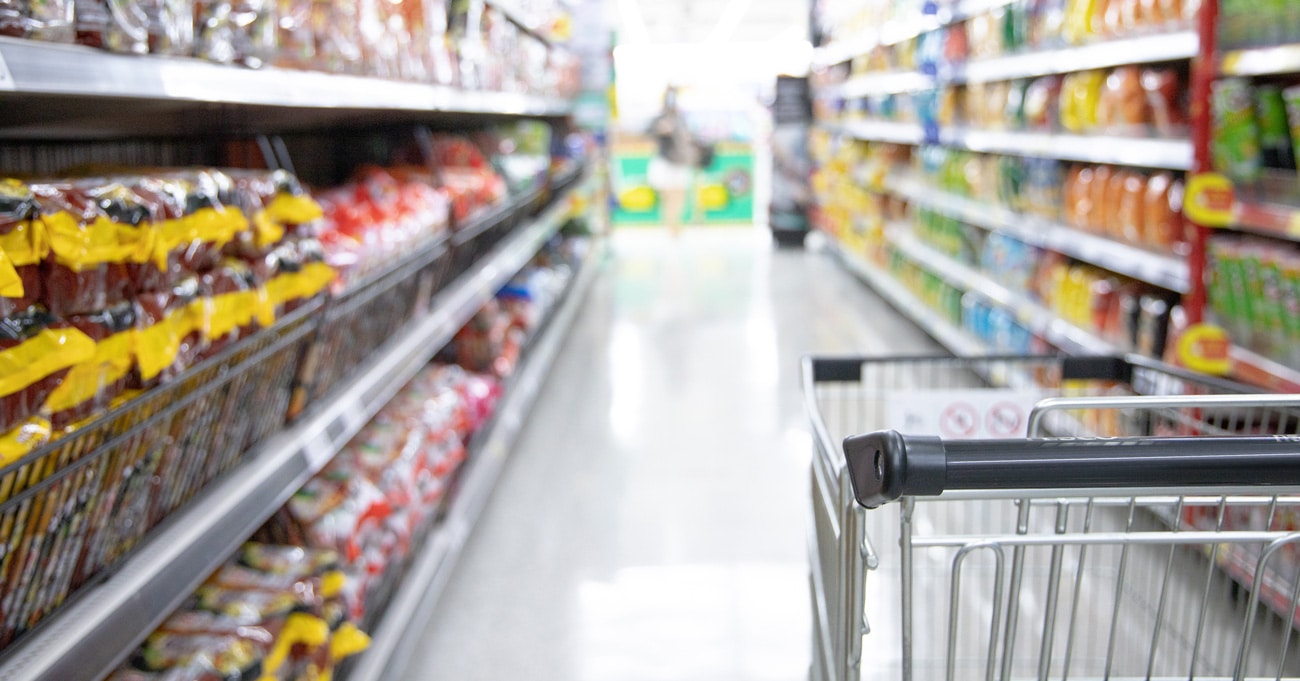 A shopping cart in a grocery store aisle, surrounded by shelves stocked with various colorful packaged food items. The image is bright and the shelves are well-organized.