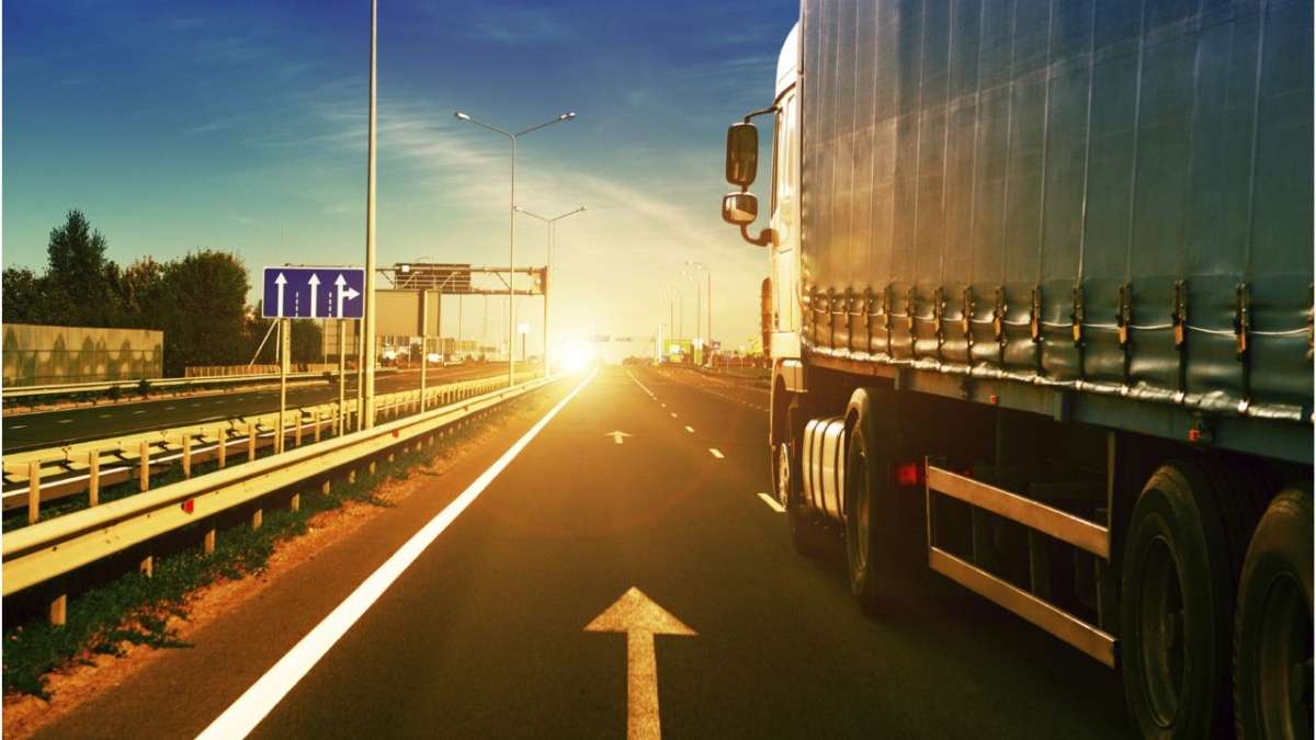 A large truck drives along a highway at sunset, with sunlight illuminating the road ahead. The highway has clear lane markings, road signs, guardrails, and a few vehicles in the distance.