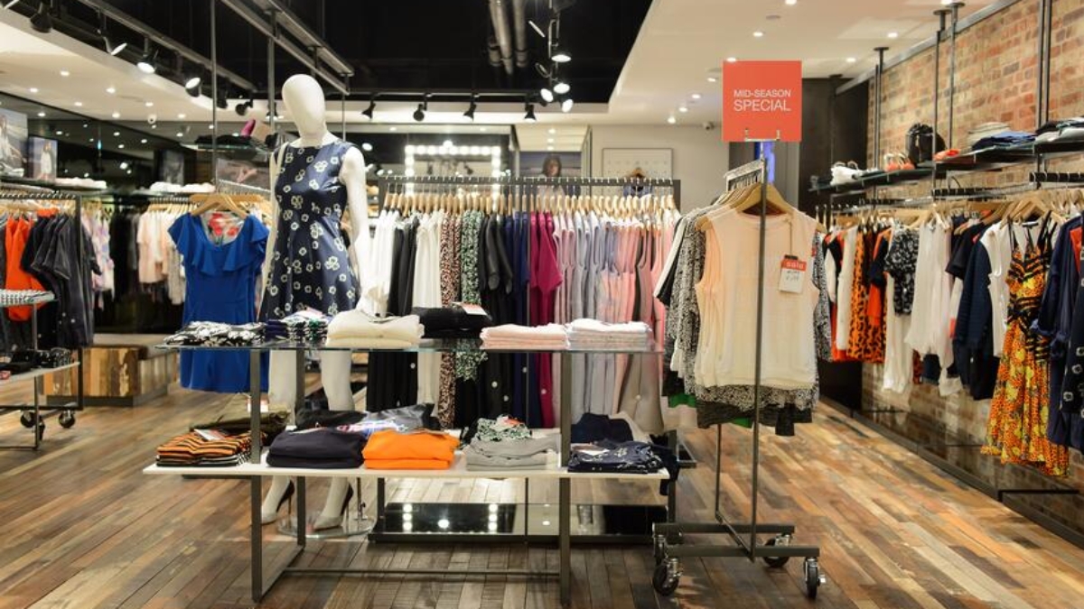 Clothing store interior with wooden floors, racks of colorful dresses and shirts, a mannequin in a blue dress, folded clothes on tables, and a sign that reads Mid-Season Special. Lean retail principles shine in the bright, modern lighting overhead.