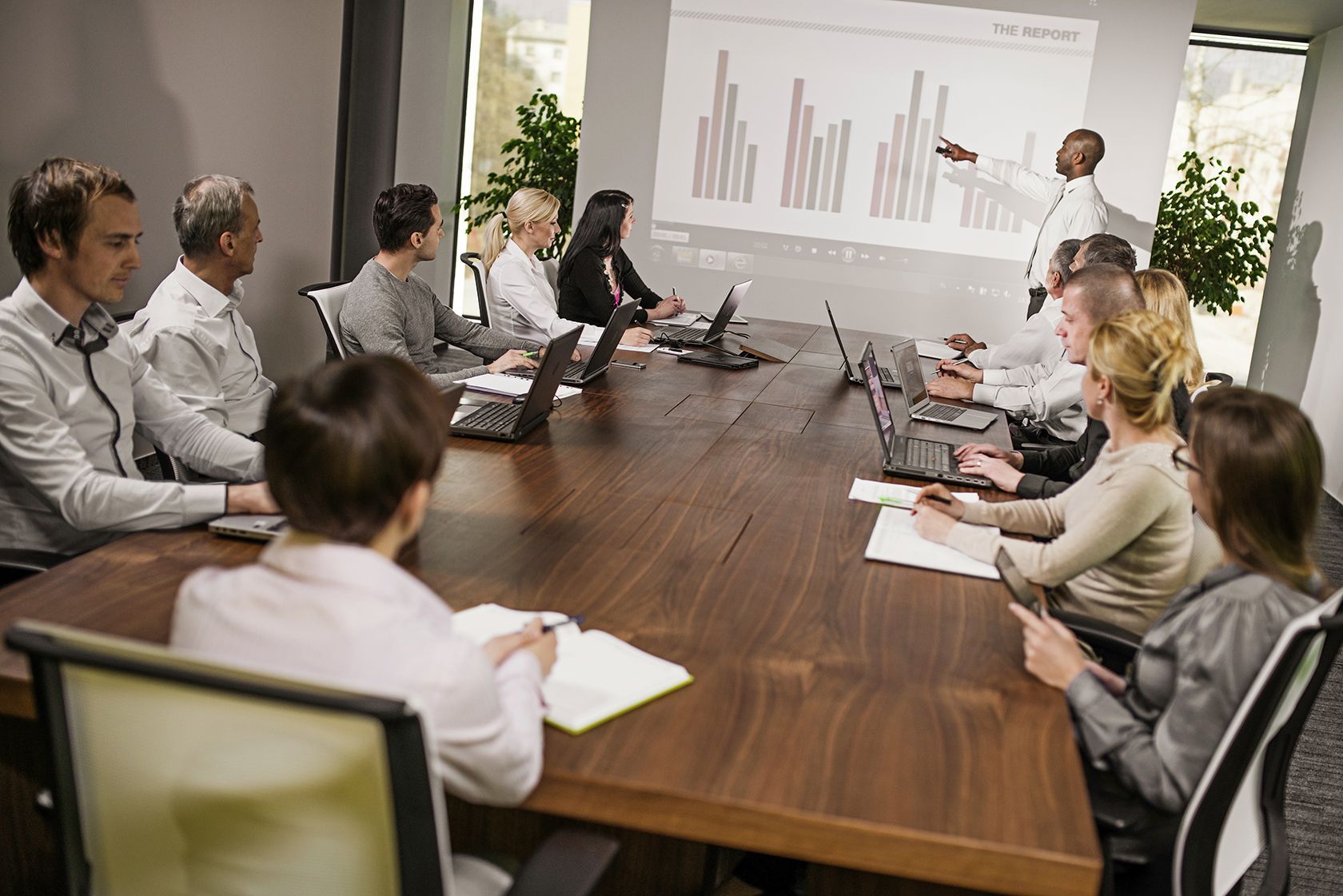 A group of people in business attire sit around a conference table with laptops and notepads, watching a presenter point to a bar chart on a projected screen during a meeting about the Microsoft Dynamics AX upgrade.
