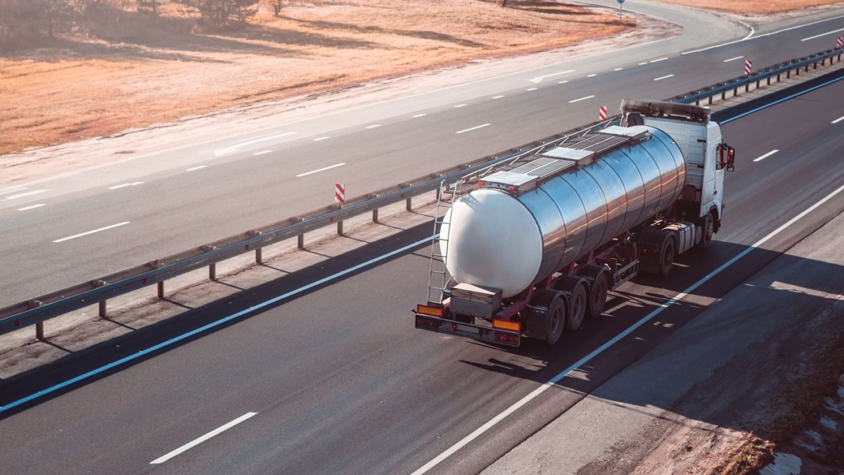 A silver tanker truck drives along an empty multi-lane highway on a clear day, with dry grass visible on the roadside.