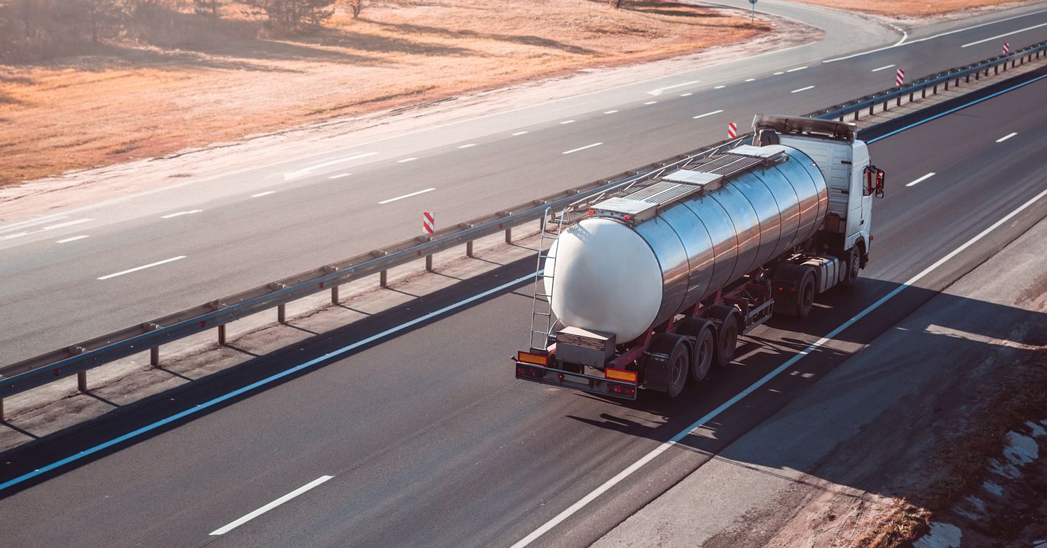 A silver tanker truck drives along an empty multi-lane highway on a clear day, with dry grass visible on the roadside.