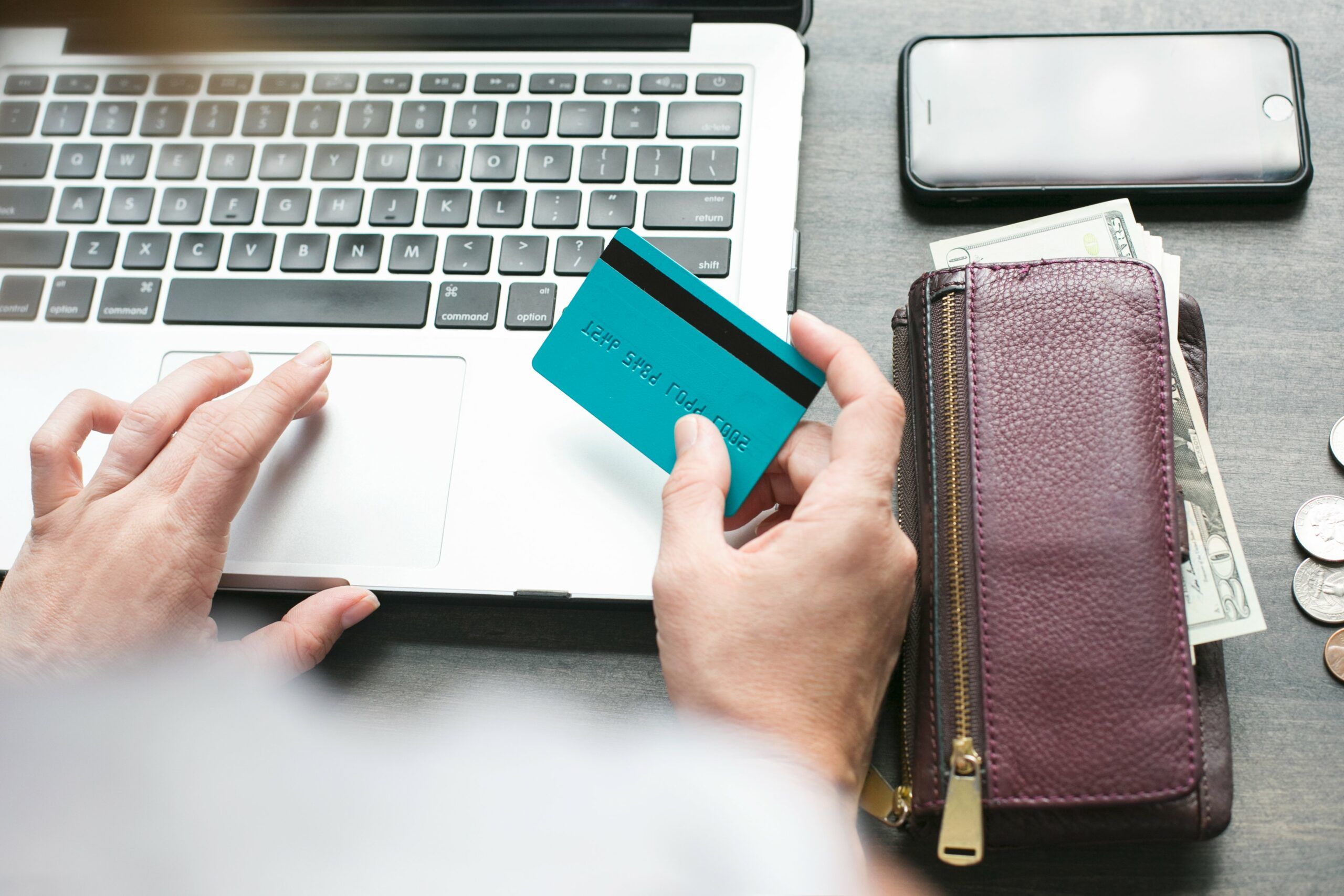 A person uses a laptop while holding a blue credit card; a purple wallet with cash, coins, and a smartphone are nearby on the table.