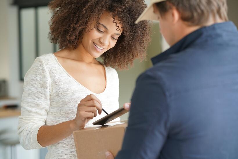 A woman smiles as she receives a package from a delivery person and signs for it on a digital device.
