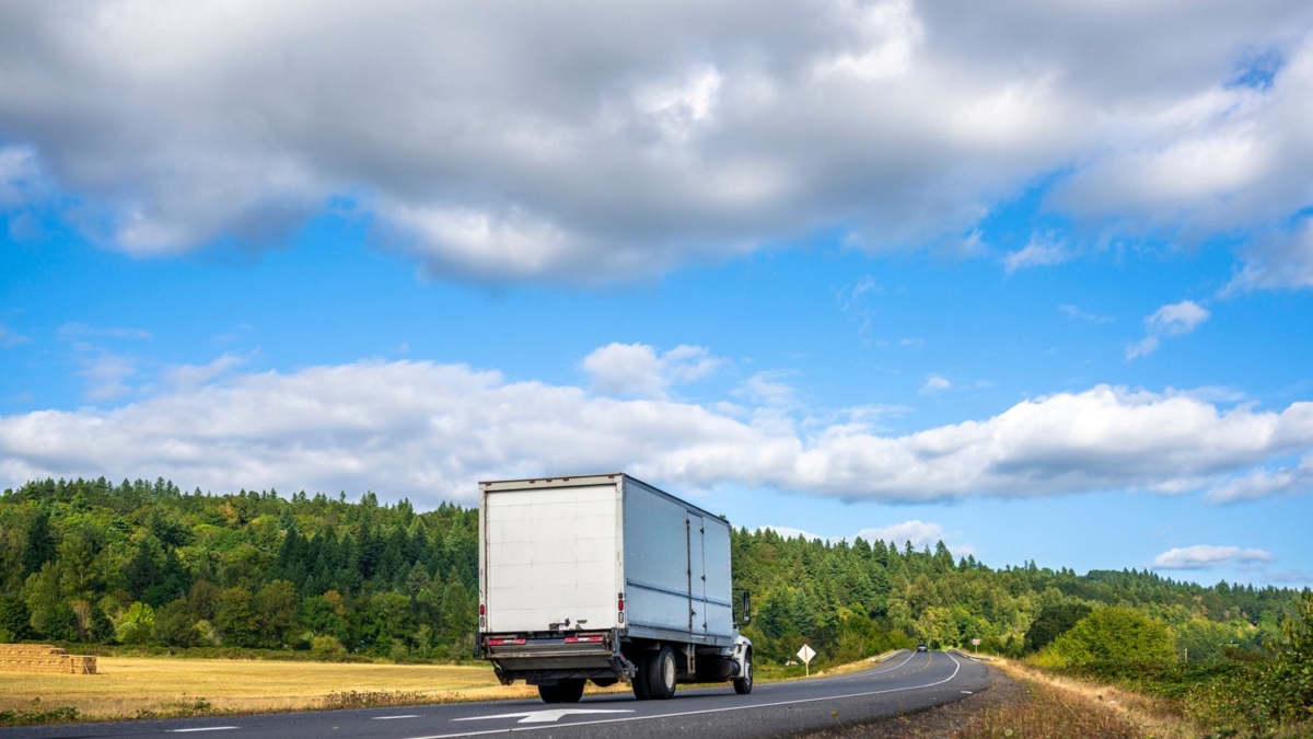 A white truck drives on a winding rural road bordered by green trees and fields, with a blue sky and scattered clouds overhead.