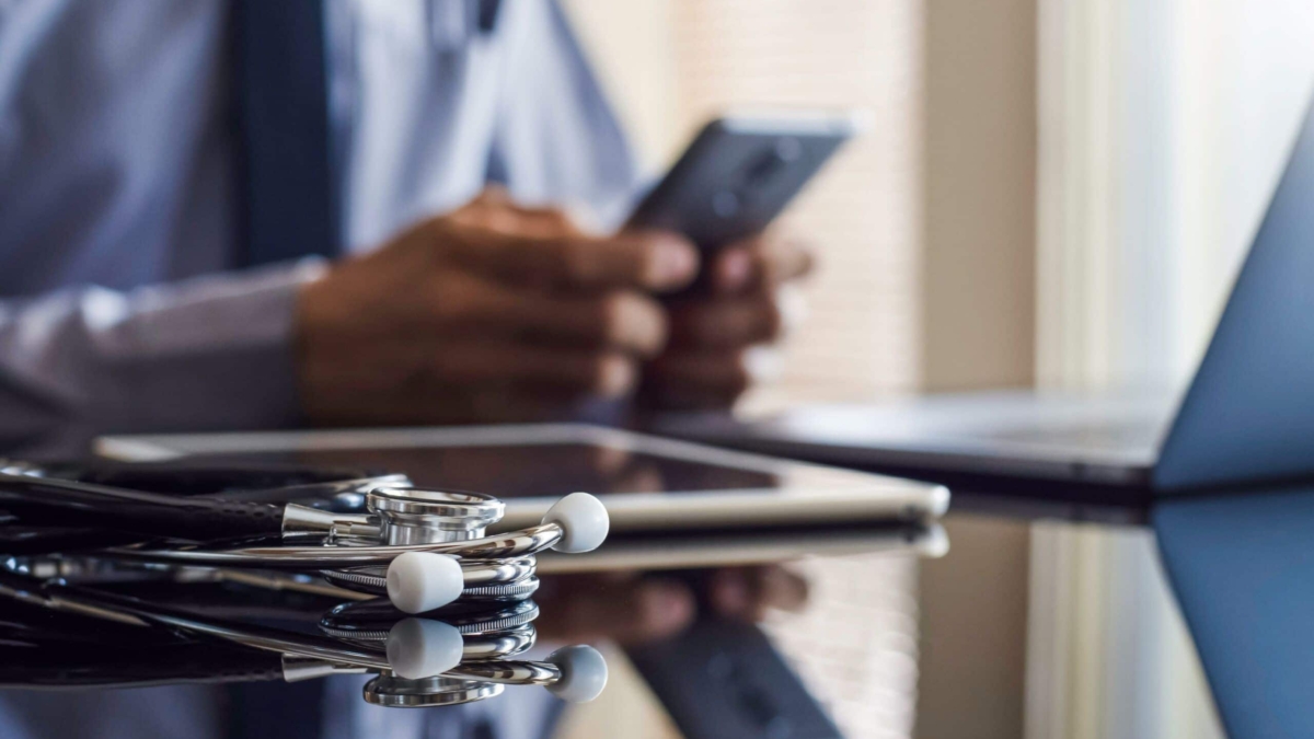 A stethoscope rests on a desk near a tablet and laptop, while a person in business attire uses a smartphone in the background, suggesting a medical or healthcare professional at work.