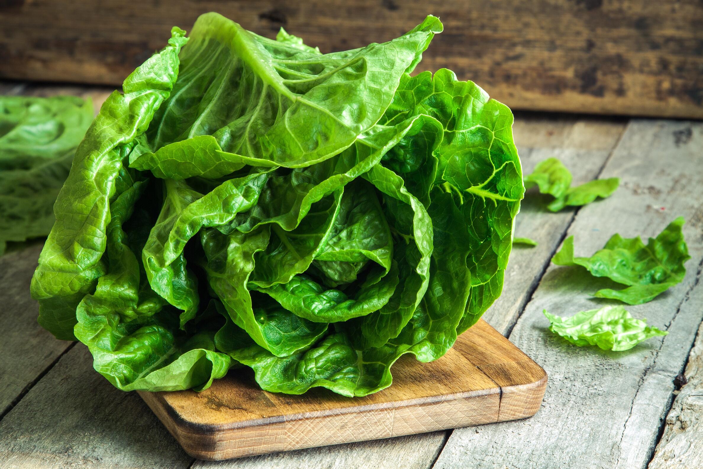 shutterstock_249162988 A fresh head of green lettuce sits on a small wooden cutting board, with a few loose lettuce leaves nearby, on a rustic wooden surface.