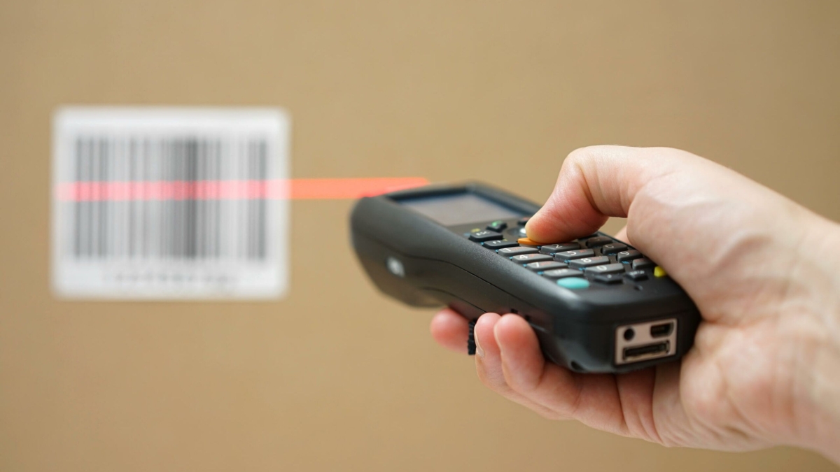 A hand holds a barcode scanner, aiming a red laser at a barcode on a cardboard box, likely for inventory or shipping purposes.