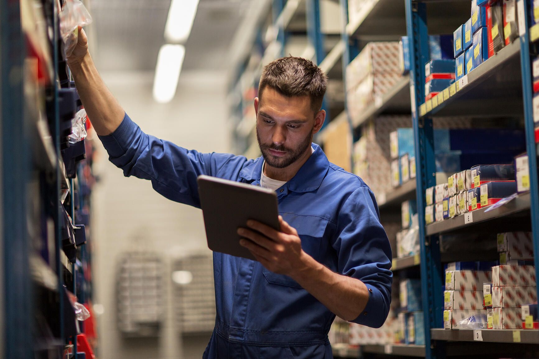 A man in a blue work jumpsuit stands between warehouse shelves, reaching for a box with one hand and holding a tablet in the other, checking inventory or stock.