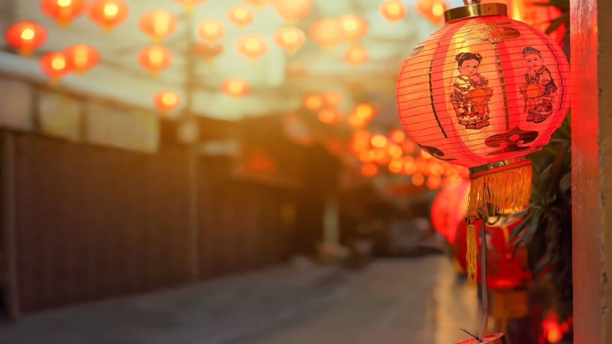 A red paper lantern with an illustration of a woman in traditional attire hangs outside, glowing among many similar lanterns lining a street in the evening. The background is softly blurred, emphasizing the lanterns’ warm light.