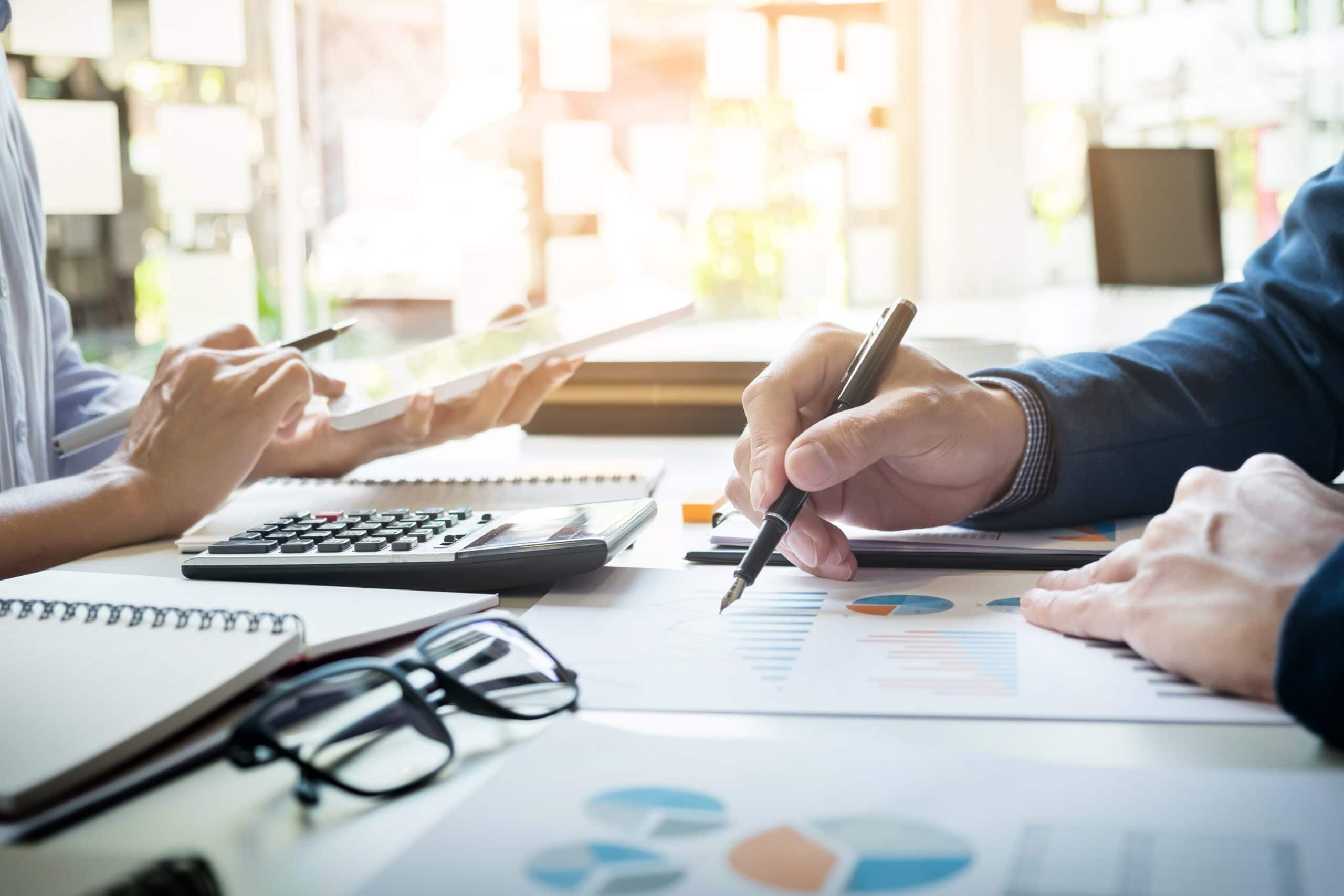 Two people working at a desk with charts, a calculator, and a tablet. One person is using a pen to point at a financial graph, while the other holds a tablet. Glasses and notebooks are also on the table.