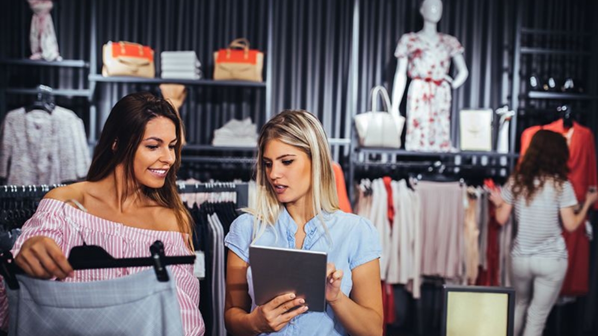 Two women are shopping for clothes in a store; one is holding a garment while the other shows her something on a tablet. Clothing and handbags are displayed in the background, with two more people browsing.