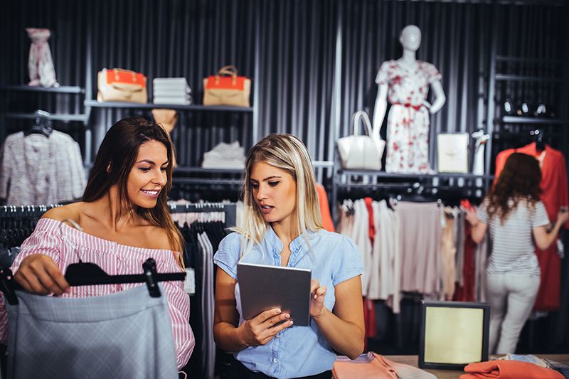 Two women stand in a clothing store, one holding a skirt and the other using a tablet. Clothing racks and accessories are visible, with another person browsing in the background. Mannequins display outfits.