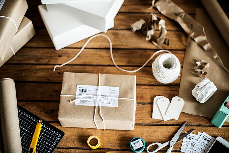 A wooden table with a parcel wrapped in brown paper and white string, surrounded by packing materials, scissors, shipping labels, blank tags, a utility knife, a ruler, tape, and white boxes.
