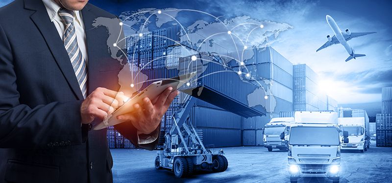 A man in a suit uses a tablet with graphics of a world map and connection lines, overlaid on a background of cargo containers, a forklift, airplanes, and trucks, representing global logistics and supply chain management.