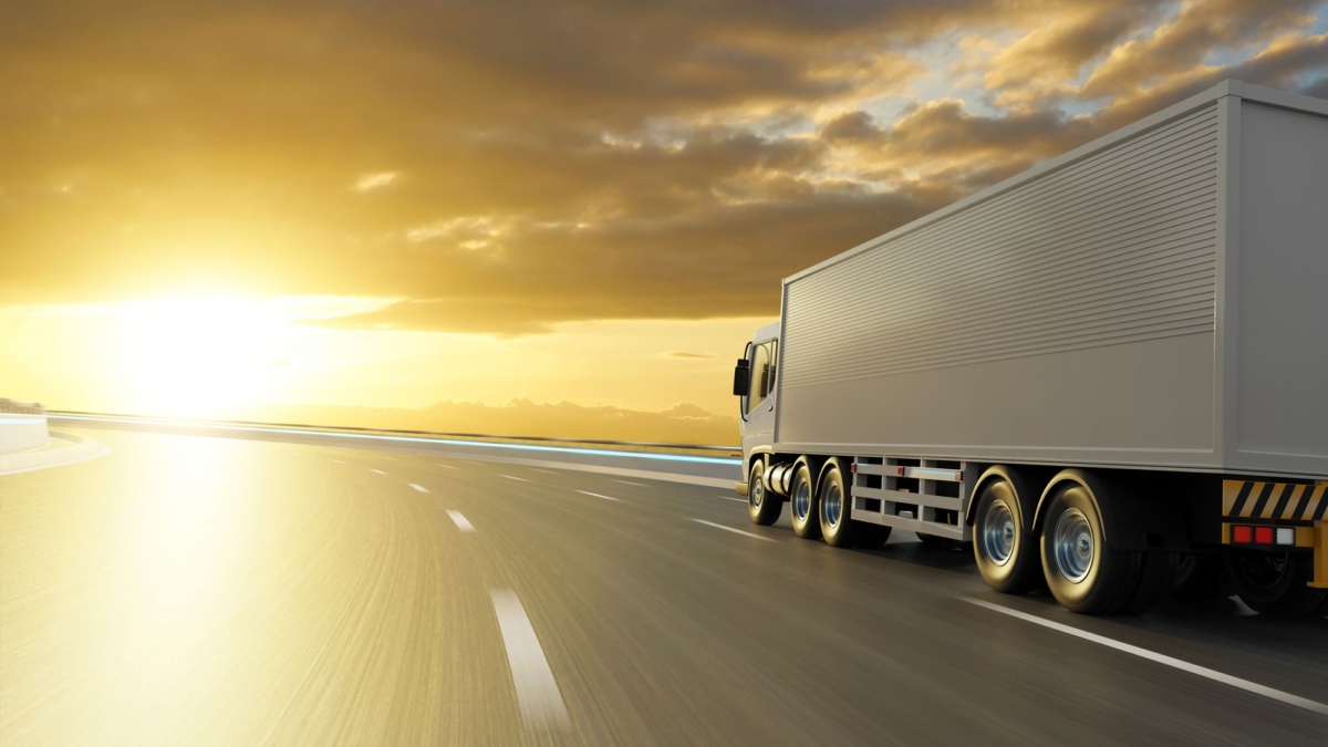 A large white semi-truck drives down an empty highway at sunset, with golden sunlight illuminating the road and dramatic clouds in the sky.