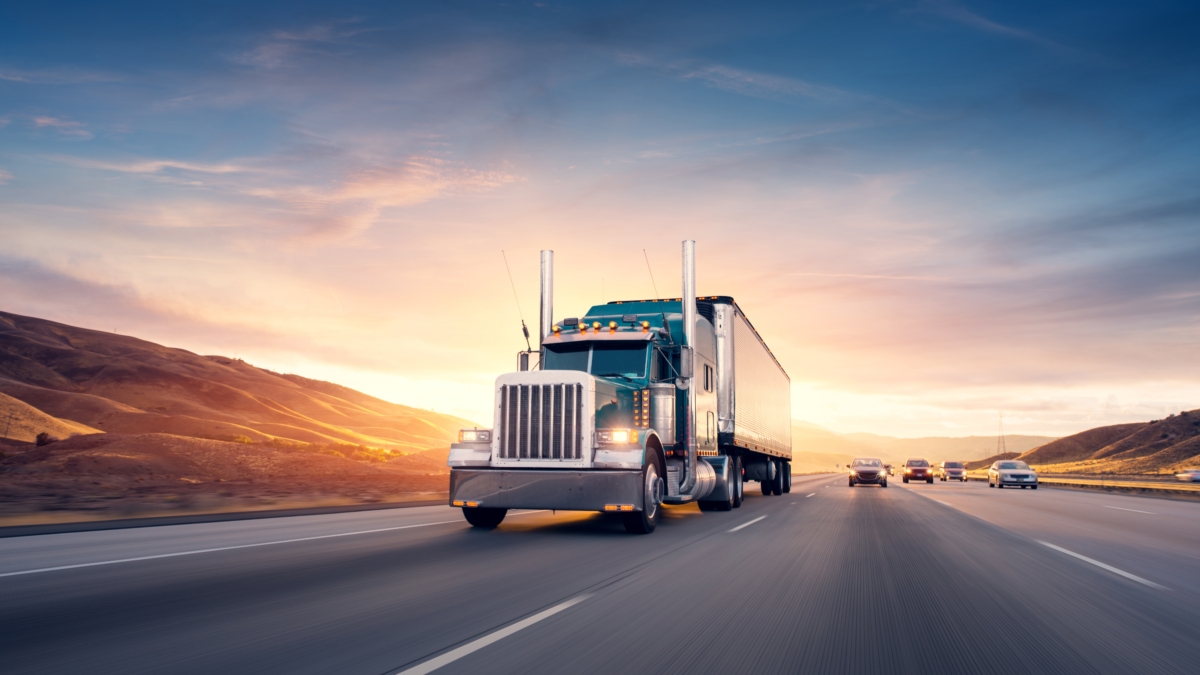 A large semi-truck drives down a highway at sunset, with rolling hills in the background and several cars traveling in the opposite direction under a colorful sky.