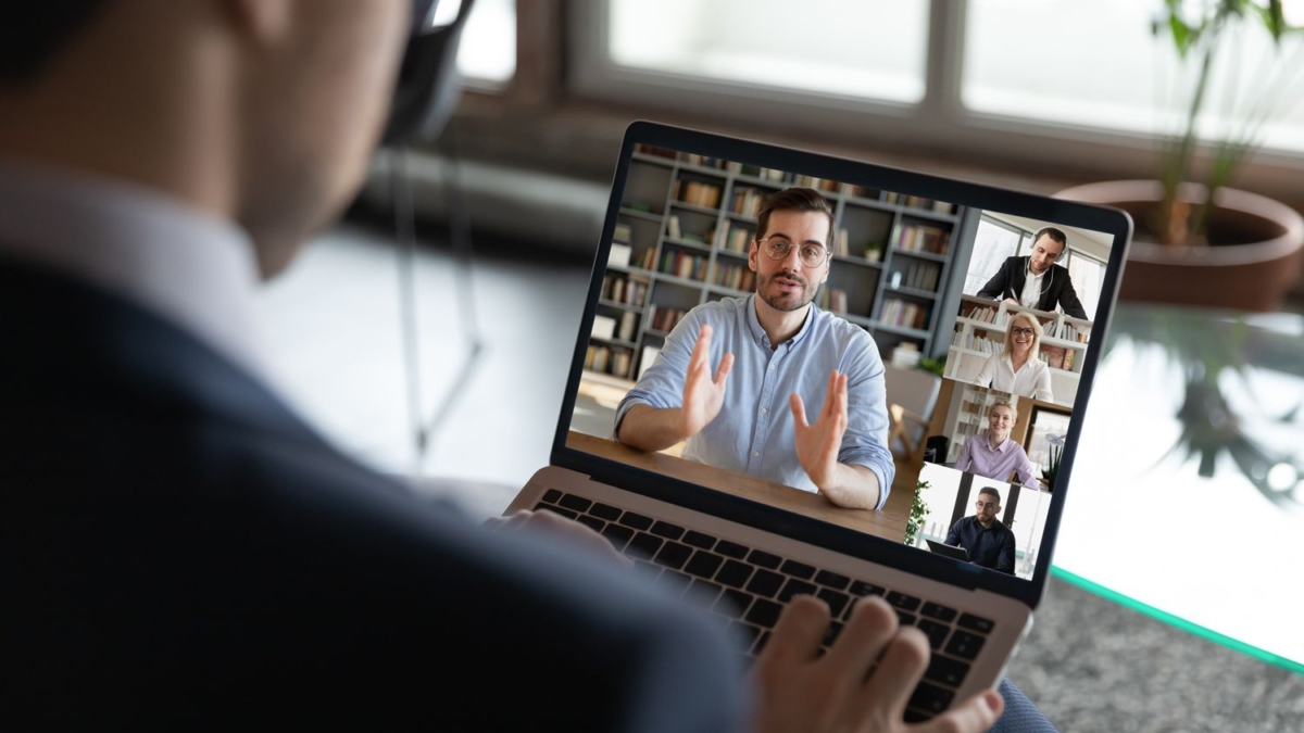 A person in a suit participates in a video conference on a laptop, showing five people on screen, each in separate windows, engaged in discussion. A potted plant and large windows are visible in the background.