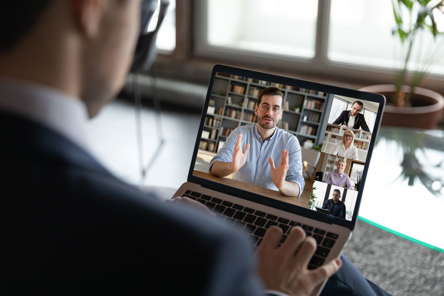 A person in a suit participates in a video conference on a laptop, showing five people on screen, each in separate windows, engaged in discussion. A potted plant and large windows are visible in the background.