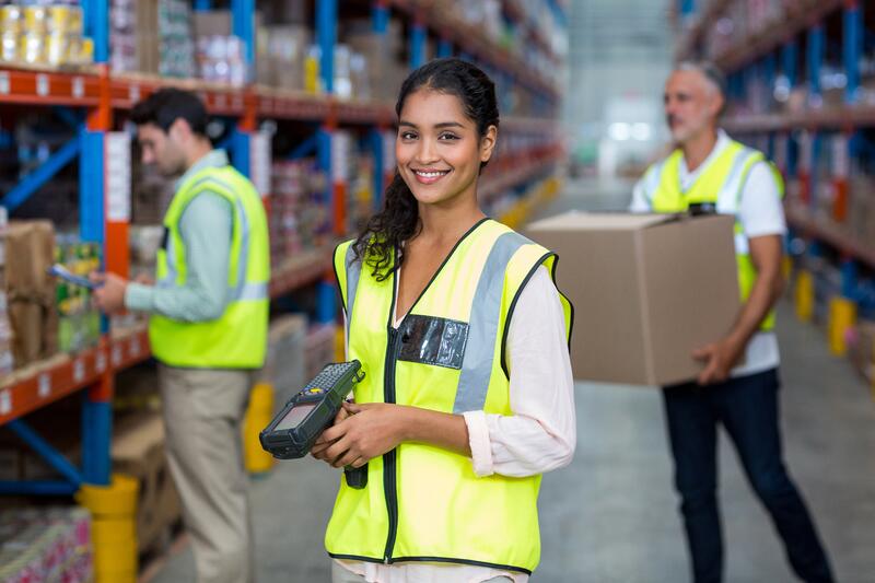 A woman in a yellow safety vest smiles at the camera while holding a barcode scanner in a warehouse. Two coworkers in safety vests work in the background, one handling a box and the other checking inventory.