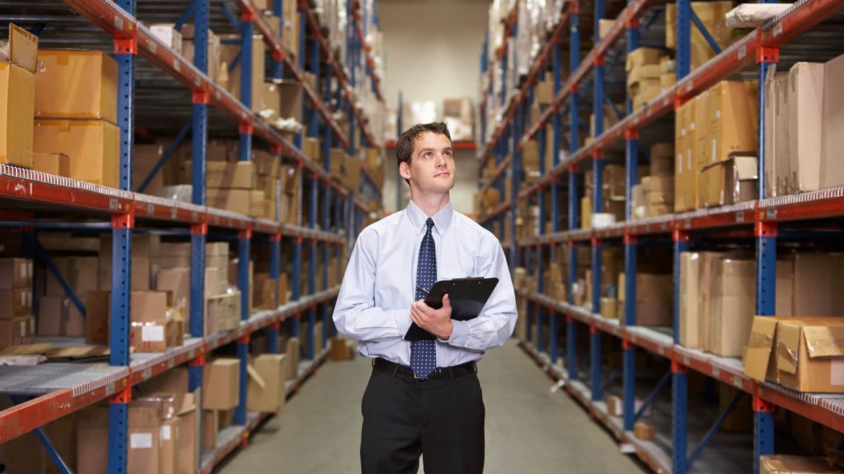 A man in business attire holding a clipboard stands in the aisle of a warehouse, surrounded by tall shelves filled with boxes and packages.