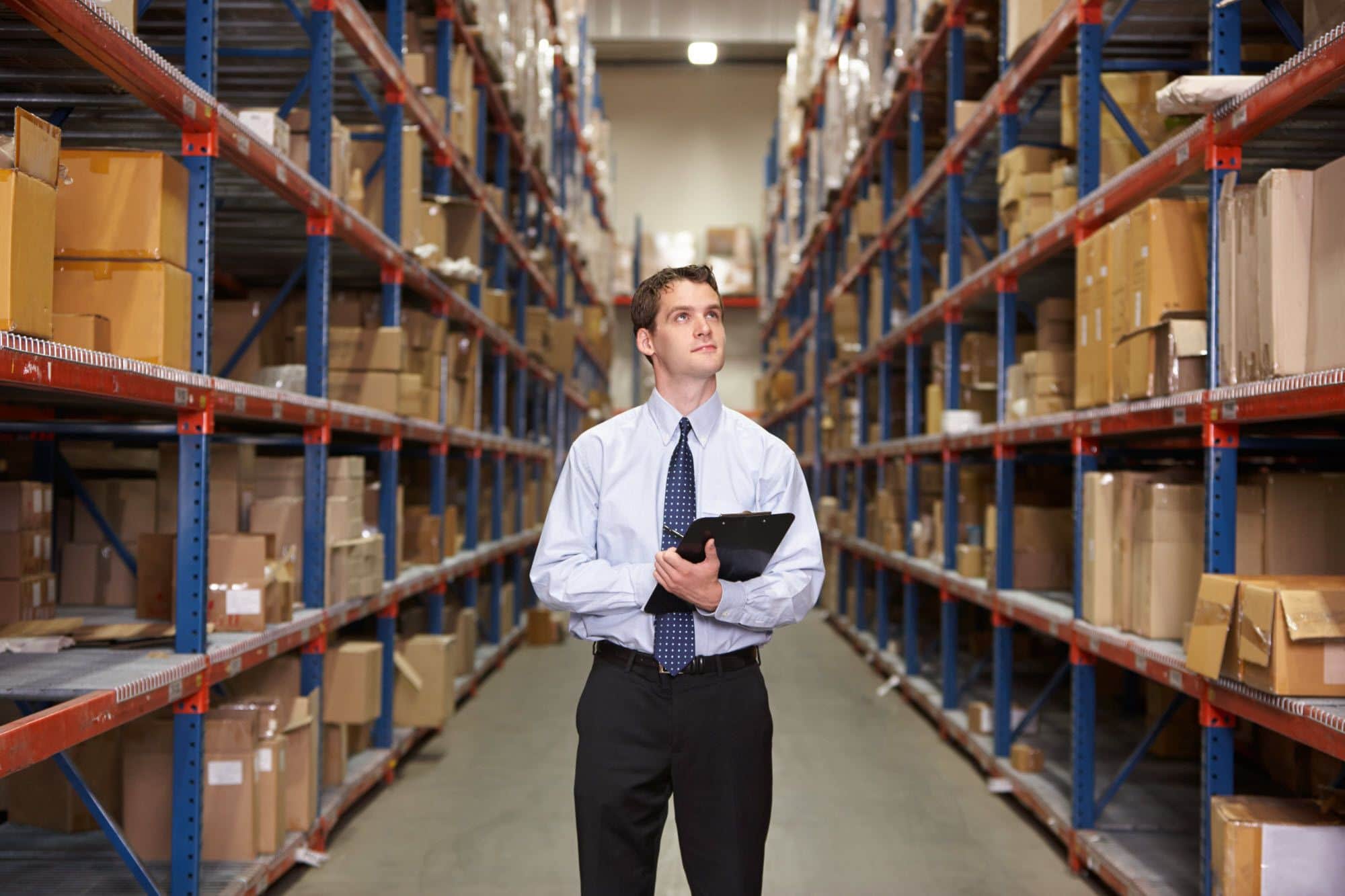 A man in business attire holding a clipboard stands in the aisle of a warehouse, surrounded by tall shelves filled with boxes and packages.