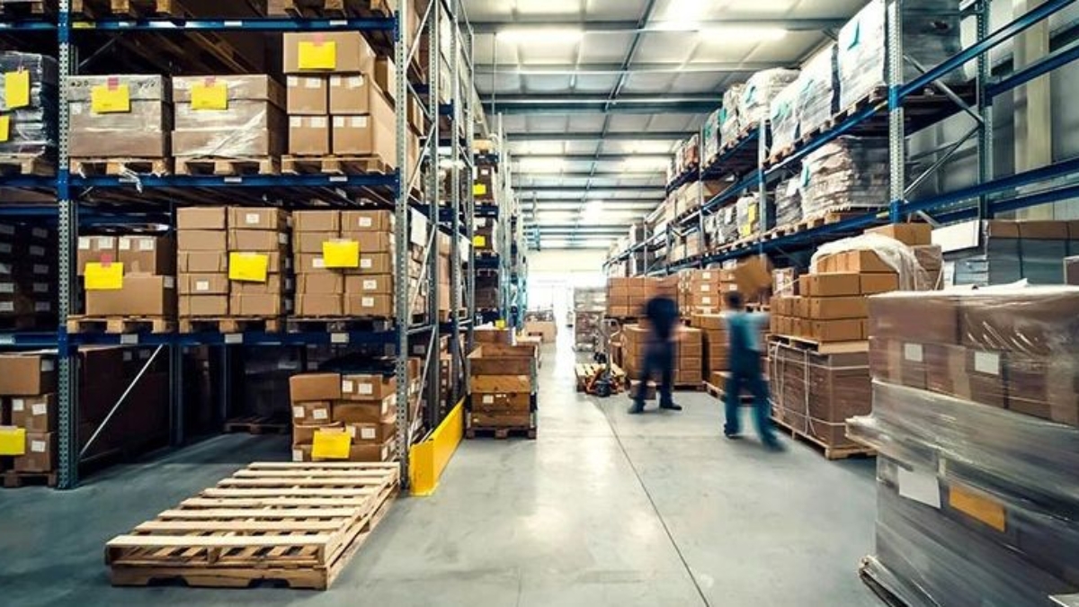 Warehouse interior with tall shelves stacked with cardboard boxes and packages. Two blurred workers, motivated by incentive based pay, are moving around, and pallets are visible in the aisle. The space is brightly lit and organized.
