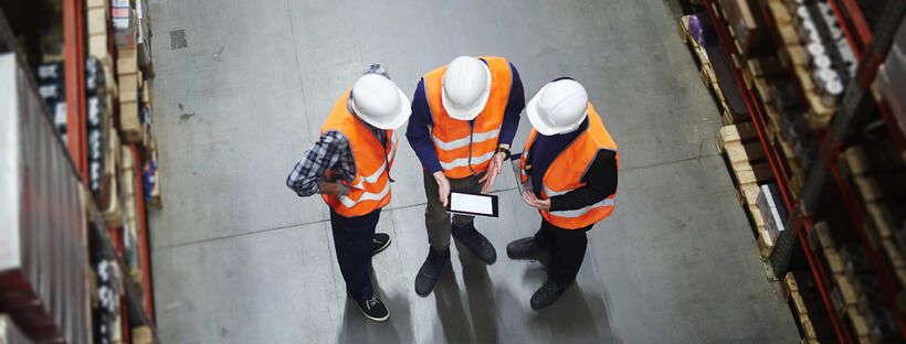 Three workers in orange safety vests and white hard hats stand in a warehouse, viewed from above, looking at a tablet together among shelves filled with boxes.