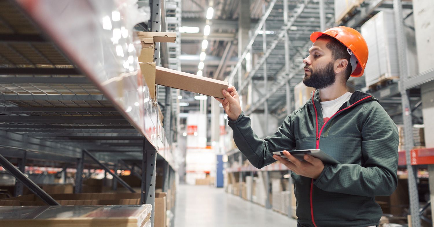 warehouse_worker (1) A man wearing an orange hard hat and dark jacket uses a tablet while placing a box on a shelf in a large warehouse with high racks and industrial lighting.