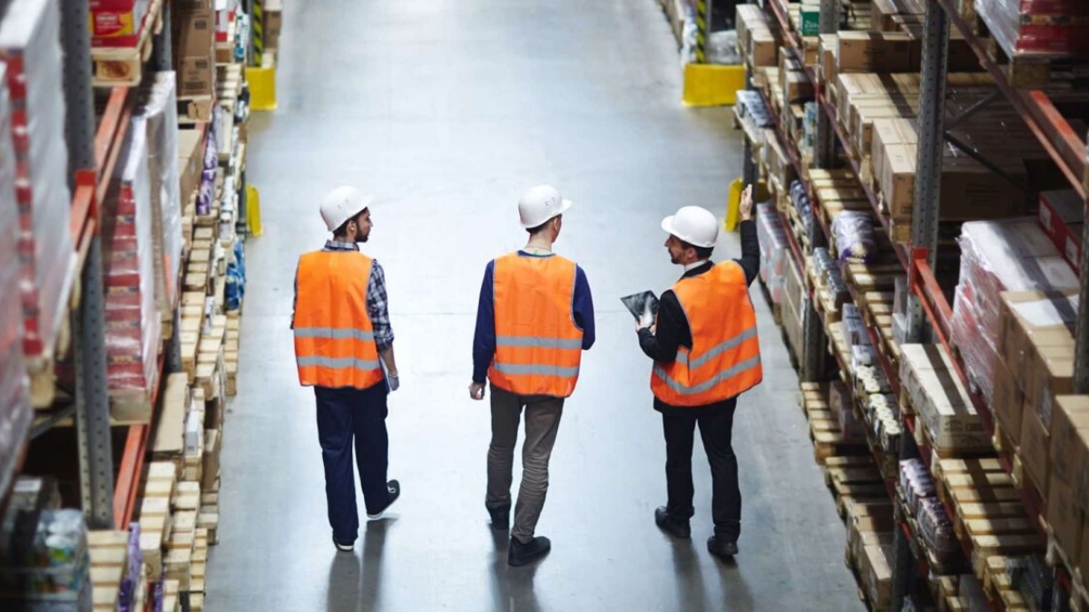 Three people wearing orange safety vests and white hard hats walk down an aisle in a large warehouse, surrounded by shelves stocked with various boxes and goods.
