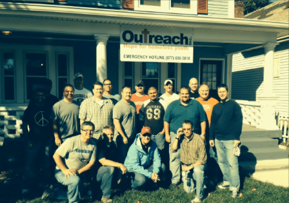Picture1 A group of fifteen men pose and smile together outside a house with a sign reading Outreach: Hope for Homeless Youth and an emergency hotline number above the porch.