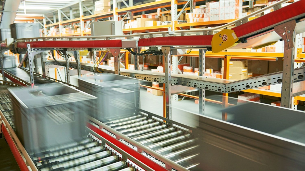 Grey storage bins move quickly along a conveyor belt system in a warehouse, with shelves stocked with various boxes and packages in the background. The motion blur highlights fast-paced automated sorting and supports efficient automation analysis.