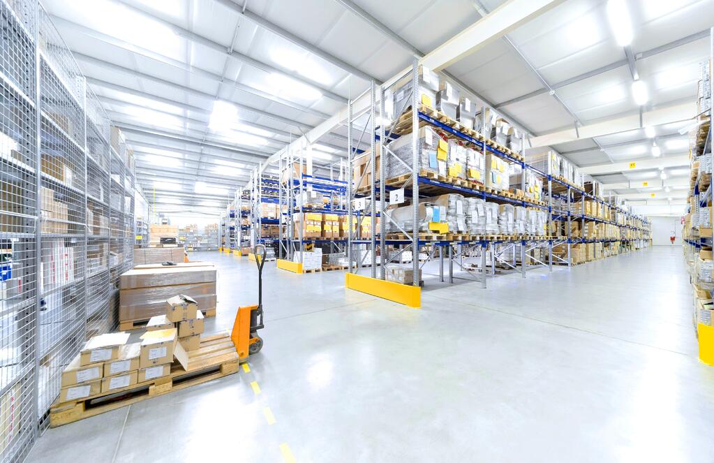 Spacious warehouse with high shelves filled with boxes and goods. Pallets and a hand pallet truck are visible on the polished concrete floor under bright overhead lighting.