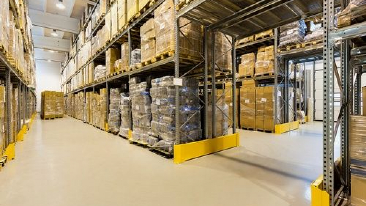 A large warehouse interior with tall metal shelves stacked with cardboard boxes and pallets. The floor is clean, and there is ample lighting. The shelves line both sides of a wide aisle.