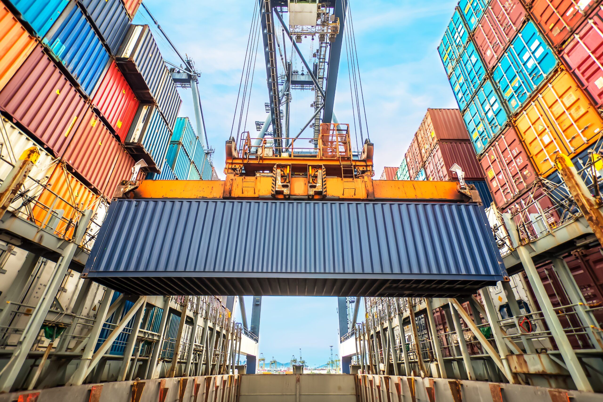 A large crane lifts a blue shipping container above a cargo ship, surrounded by stacks of colorful containers under a bright sky at a busy port.