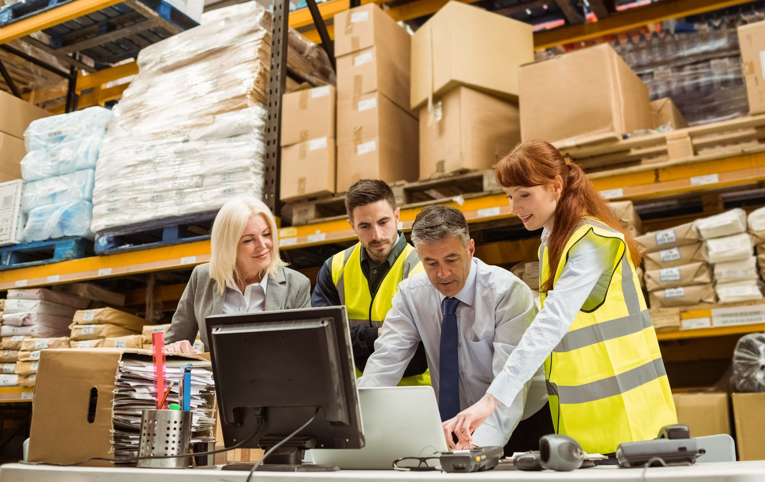 Four warehouse workers in safety vests gather around a computer and laptop, discussing work. They are surrounded by shelves filled with stacked boxes and packages in a busy warehouse environment.