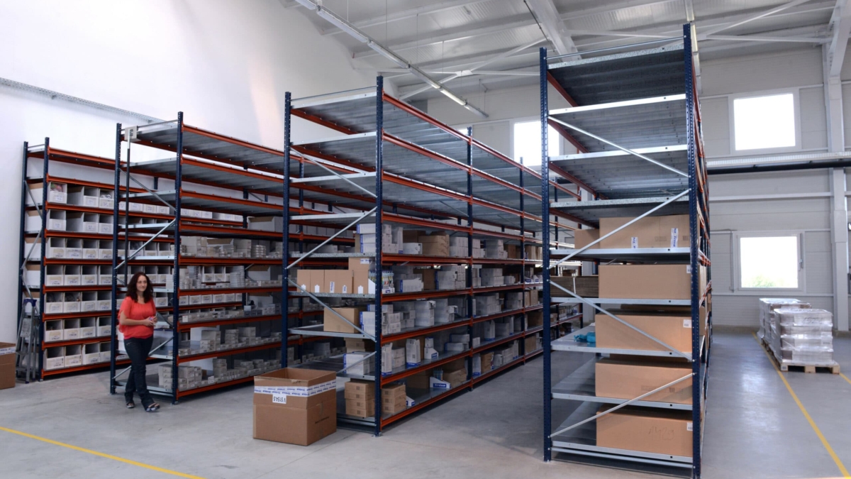 A woman walks through a spacious warehouse with tall metal shelves filled with boxes and packages. The floor is clean and there are additional boxes on the ground near the shelves.