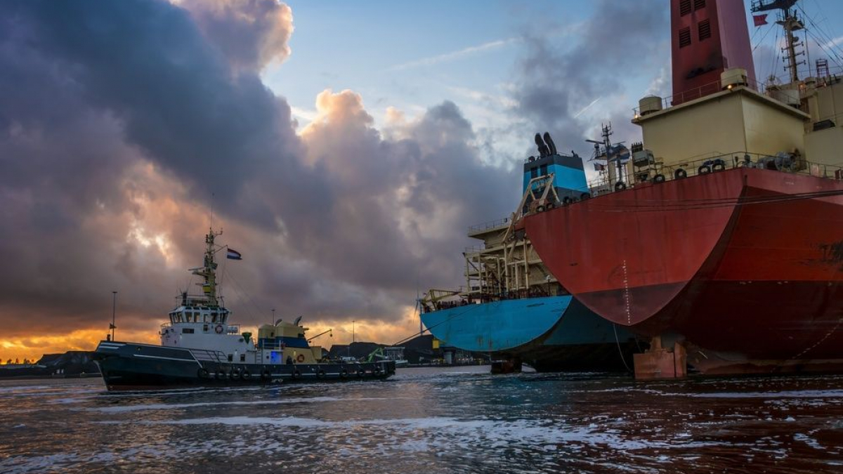 A tugboat moves near two large docked cargo ships at sunset, with dramatic clouds and golden light reflected on the water.