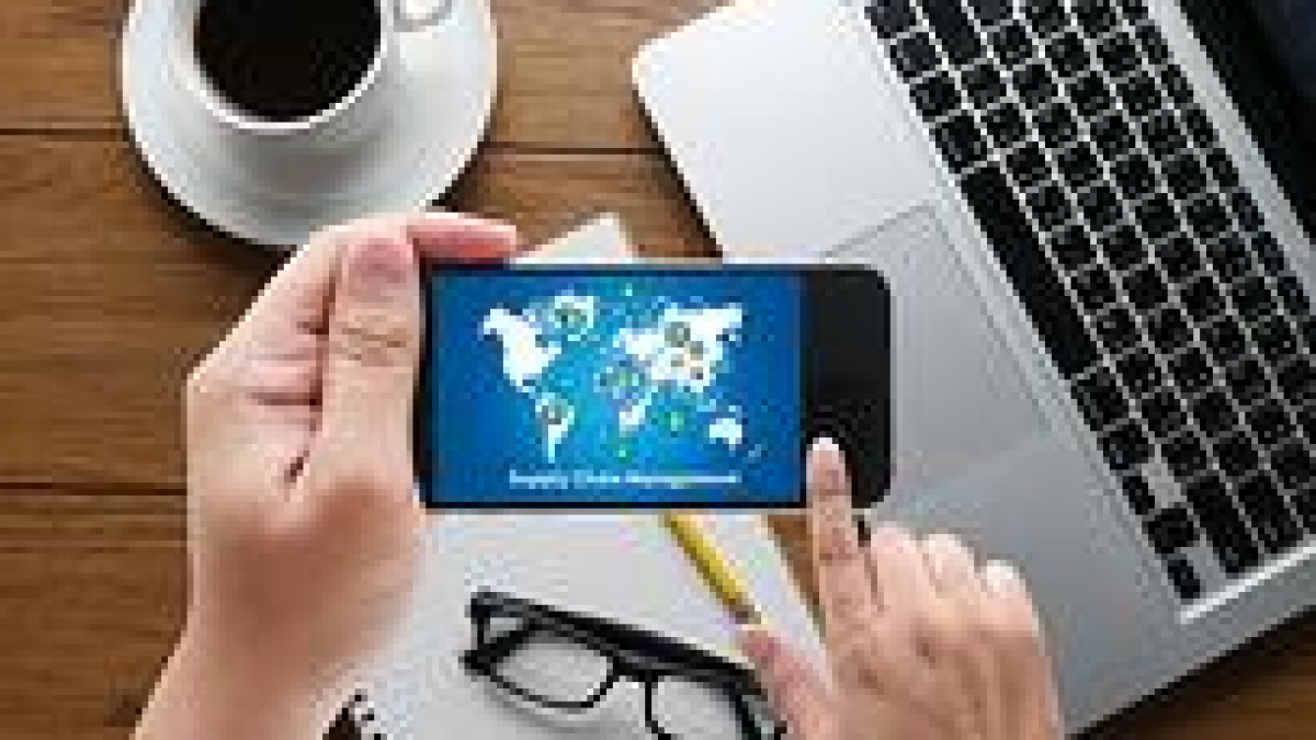 A person holds a smartphone displaying a world map with network icons. The phone is above a notepad, eyeglasses, and a keyboard, with a cup of coffee nearby on a wooden desk.