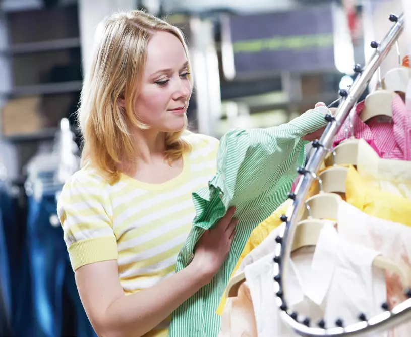 A woman with blonde hair wearing a yellow striped shirt examines a green striped shirt on a rack of colorful clothes in a store, showcasing the benefits of Multi Echelon Inventory Optimization for efficient stock management.