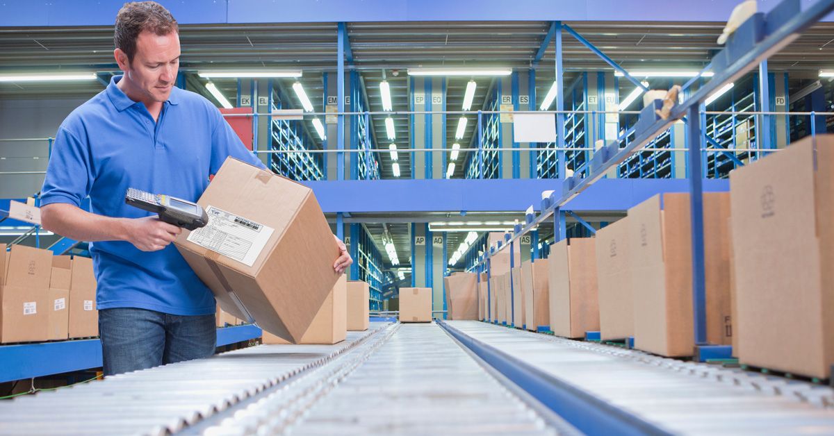A man in a blue shirt scans a barcode on a cardboard box in a large warehouse, showcasing distributed order management as he stands beside a conveyor belt lined with other boxes.