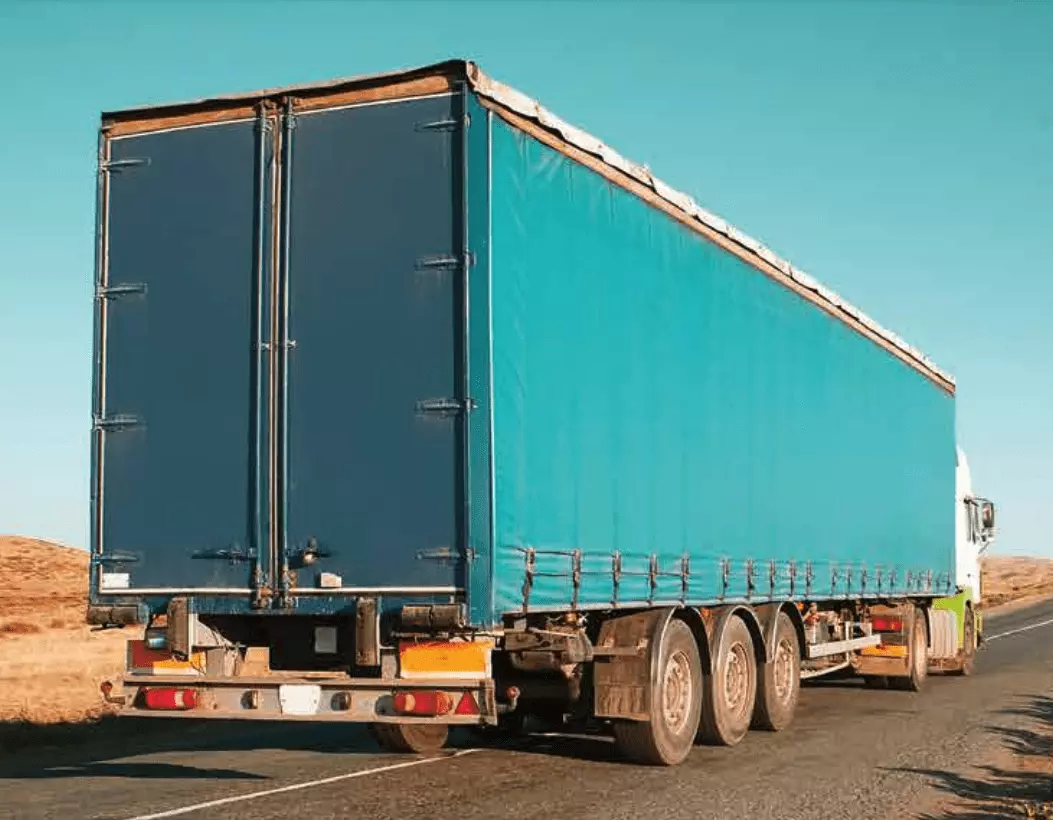 A large blue cargo truck with a covered trailer is driving on a paved road through a dry, open landscape under a clear blue sky.