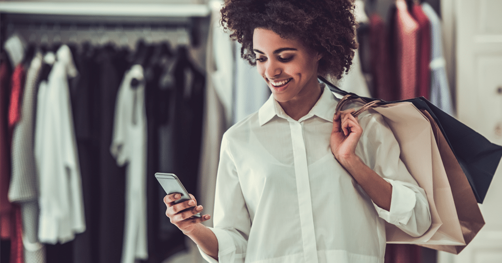 A woman holding shopping bags smiles while looking at her smartphone in a clothing store, with racks of clothes visible in the background.